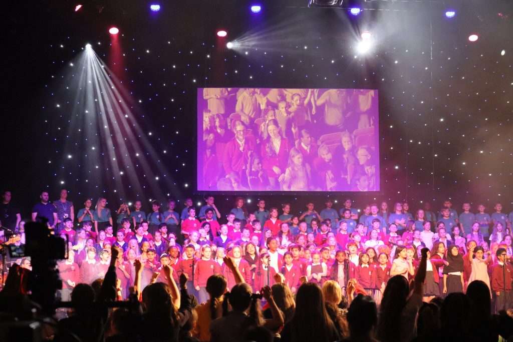 A photo of the Voices Together event with a large group of school children from a variety of schools on stage.